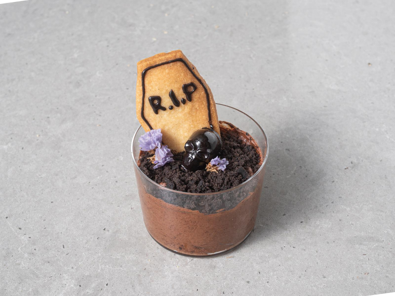 Wegański czekoladowy mus z aquafaby z pokruszonym czarnym ciastem, czekoladową czaszką i ciasteczkiem w kształcie trumny oraz kwiatami w szkalneczce.