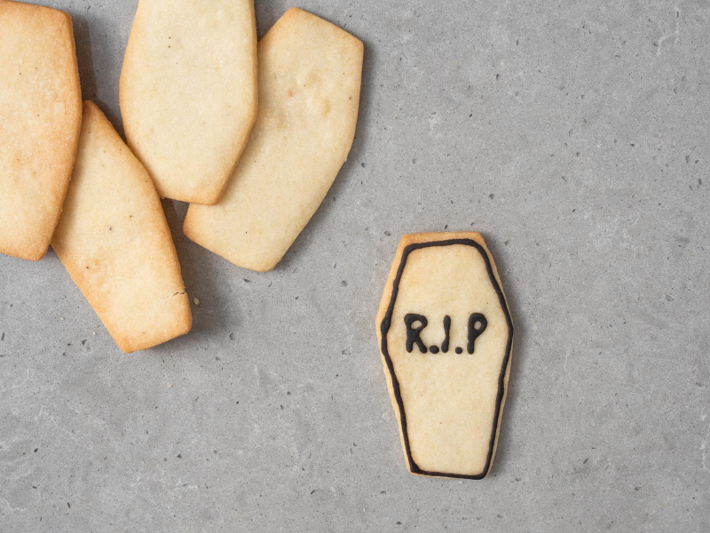 Udekorowane ciasteczko w kształcie trumny z napisem R.I.P. Obok więcej ciasteczek.,