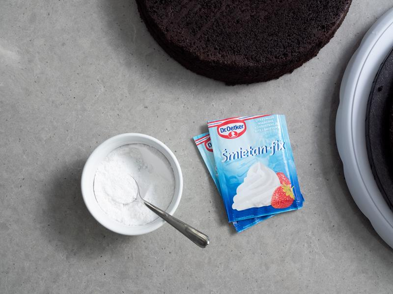 miseczka z cukrem pudrem wymieszanym ze śmietan-fixem, obok dwa opakowania śmietan-fixu oraz blaty biszkoptu