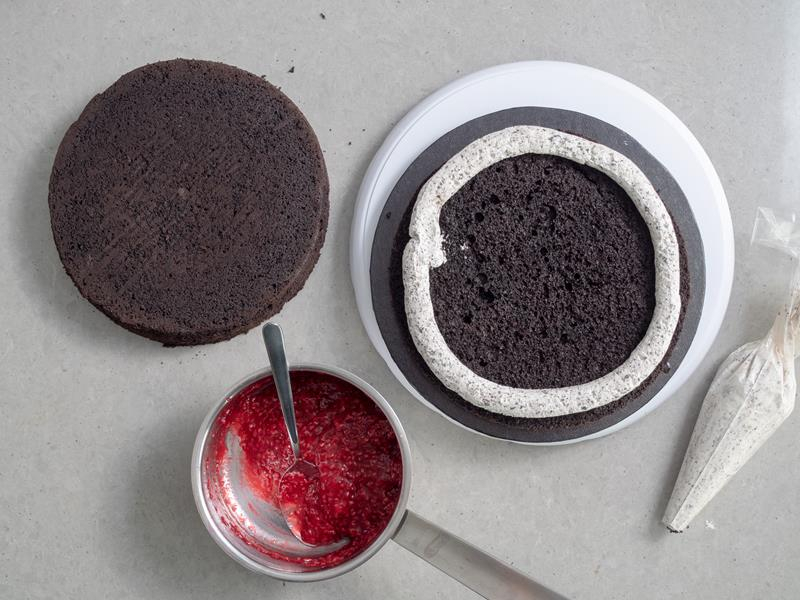 krem śmietankowy z ciasteczkami oreo w rękawie cukierniczym oraz zrobiony z kremu okrąg na brzegu jednego z blatów biszkoptu, obok rondelek z frużeliną malinową oraz drugi blat biszkoptu