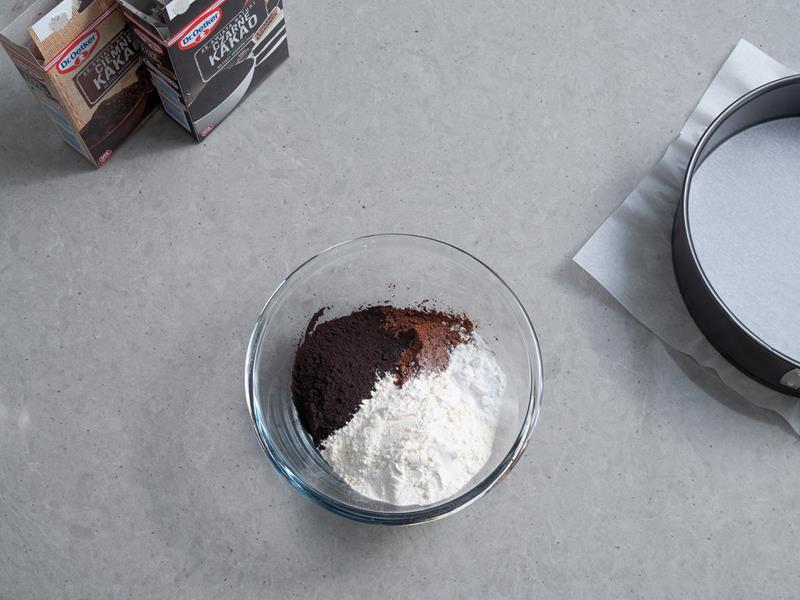 szklana miseczka z mąką pszenną kakao ciemnym, kakao czarnym oraz proszkiem do pieczenia, obok opakowanie kakao ciemnego dr. oetkera, opakowanie kakao czarnego intense dr. oetkera oraz tortownica black trend dr. oetkera wyłożona papierem do pieczenia