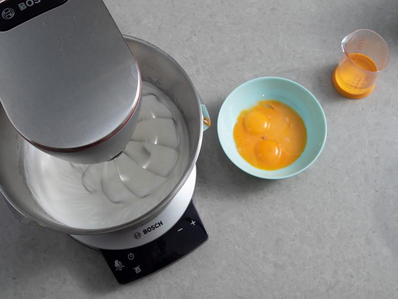 białka ubite z cukrem w misie robota kuchennego, obok miseczka z żółtkami oraz mała miarka kuchenna z olejem