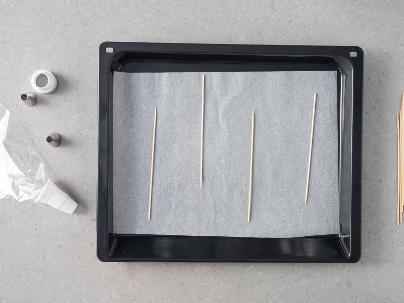 Blacha z wyposażenia piekarnika wyłożona papierem do pieczenia z drewnianymi patyczkami. do szaszłyków, obok tylka i rękaw cukierniczy