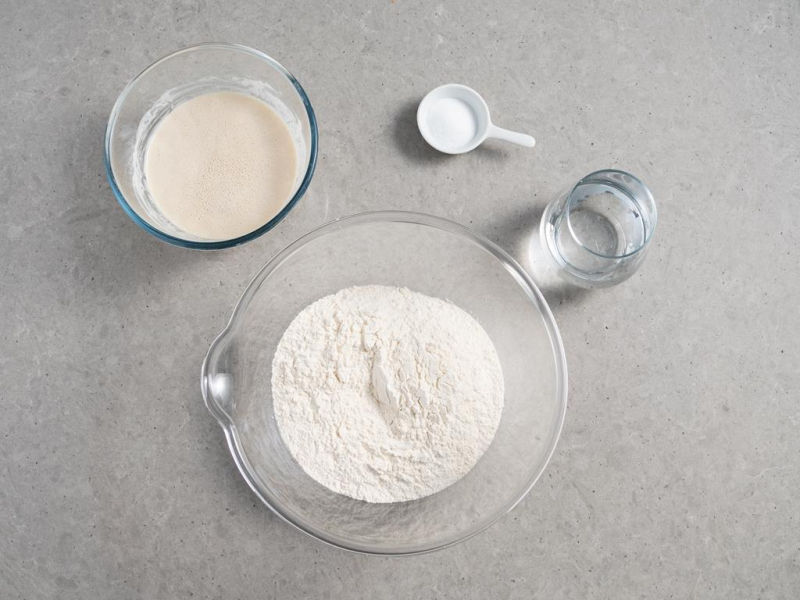 mąka pszenna w misce, zaczyn w drugiej misce, szklanka z wodą i sól na łyżeczce