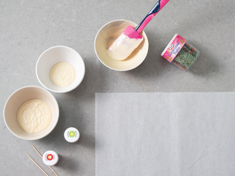 W trzech miseczkach rozpuszczona biała czekolada. Obok barwnik czerwony i zielony firmy Wilton i dekoracje cukrowe "Ciasteczkowa bajka" Dr. Oetkera