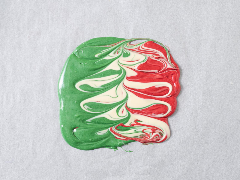 rozlana na papierze do pieczenia czekolada w trzech kolorach: zielonym, białym i czerwonym, i wymieszana za pomocą patyczka do szaszłyków.