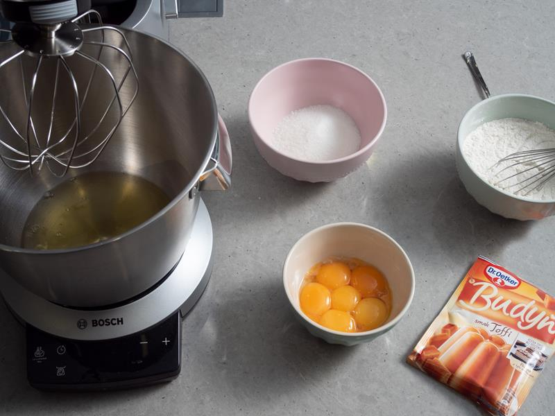 białka jaj w misie robota kuchennego marki bosch, obok żółtka, cukier i mąka pszenna w osobnych miseczkach oraz opakowanie budyniu o smaku toffi dr. oetkera
