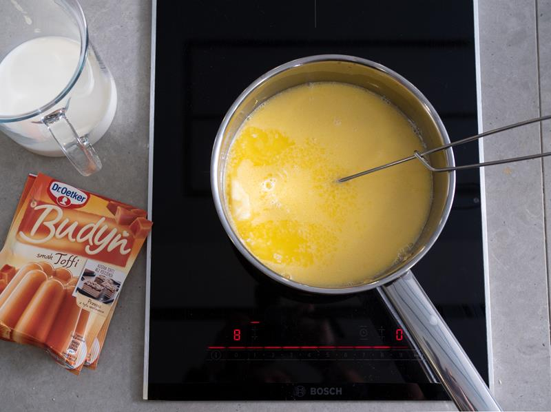 na kuchence indukcyjnej bosch rondelek z mlekiem i roztapianym w nim masłem, obok szklanka z zimnym mlekiem i dwa opakowania budyniu toffi dr. oetkera