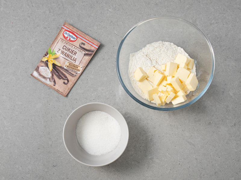 mąka i pokrojone w kostkę masło w szklanej misce, obok miseczka z cukrem i opakowanie cukru z wanilią dr. oetkera