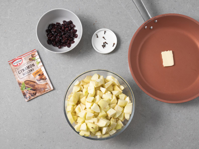 Na patelni kawałek masła. W misie pokrojone w małą kostkę jabłka. Obok żurawina suszona w miseczce, goździki na małym talerzyku i opakowanie cynamonu mielonego Dr. Oetkera