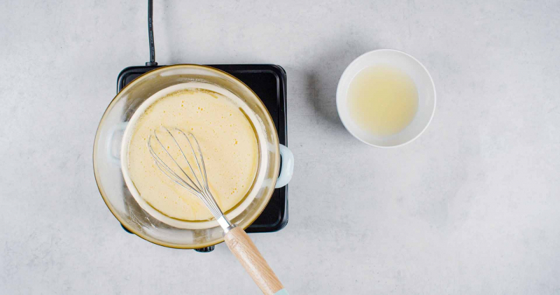 Jajko, żółtka i cukier w szklanej misce z rózgą kuchenną, obok roztopione masło i sok z cytryn z osobnych miseczkach