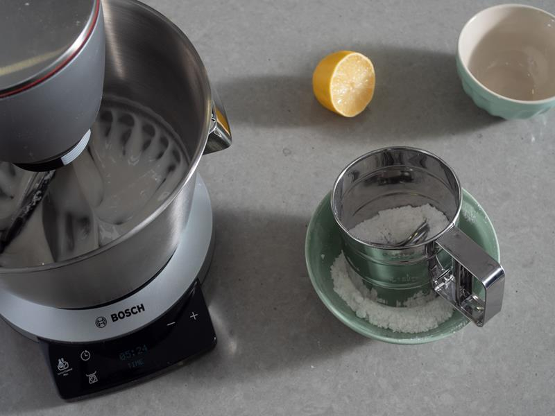 robot kuchenny bosch, w misie lukier królewski, miseczka z cukrem pudrem i przesiewaczem, połówka cytryny i pusta miseczka