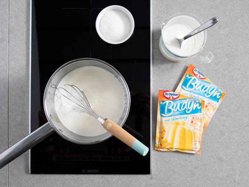 przygotowanie budyniu - rondelek z mlekiem na palniku, miseczka z cukrem, naczynie z mlekiem i rozmieszanym budyniem oraz dwa opakowania budyniu o smaku waniliowym od dr. oetkera