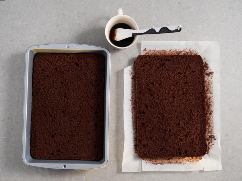 biszkopt kakaowy z olejem przekrojony na dwa blaty, jeden z blatów umieszczony z powrotem w formie do pieczenia, obok kubek z kawą i pędzelek do nasączenia blatów