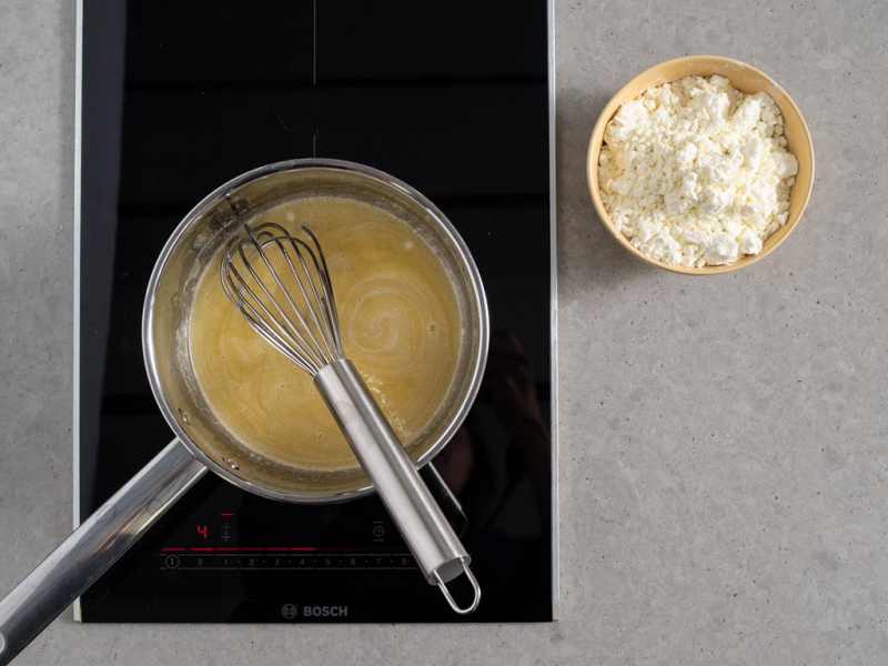 na palniku kuchenki rondelek z rozpuszczonym masłem cukrem, wodą i ekstraktem z wanilii oraz rózgą kuchenną, obok miseczka z mlekiem w proszku