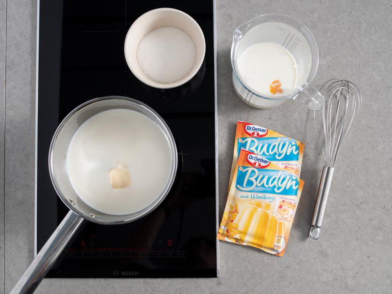mleko z masłem w rondelku na palniku kuchenki, cukier w miseczce, mleko z żółtkami w pojemniku, dwa opakowania budyniu waniliowego Dr. Oetkera oraz rózga kuchenna