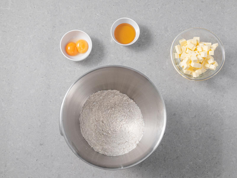 Mąka pszenna w misie z proszkiem do pieczenia i cukrem pudrem. Obok w misie zimne masło, żółtka i miód.
