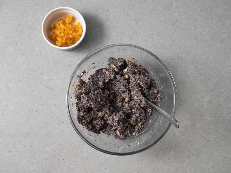 Mak wymieszany z bakaliami w szklanej misce, obok miseczka z kandyzowaną skórką z pomarańczy.