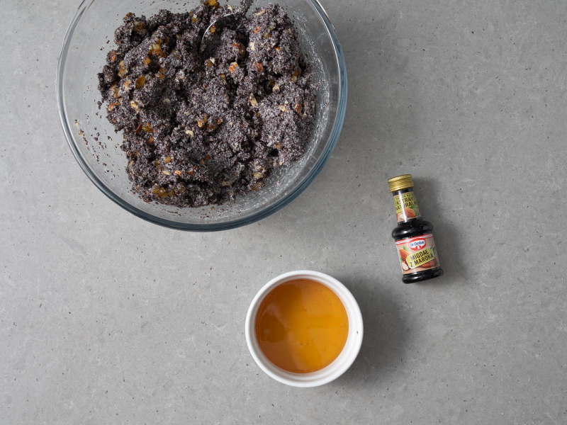 Mak wymieszany z bakaliami i skórką z pomarańczy w szklanej misce. Obok w miseczce miód oraz opakowanie ekstraktu migdałowego migdał z maroka dr. oetkera