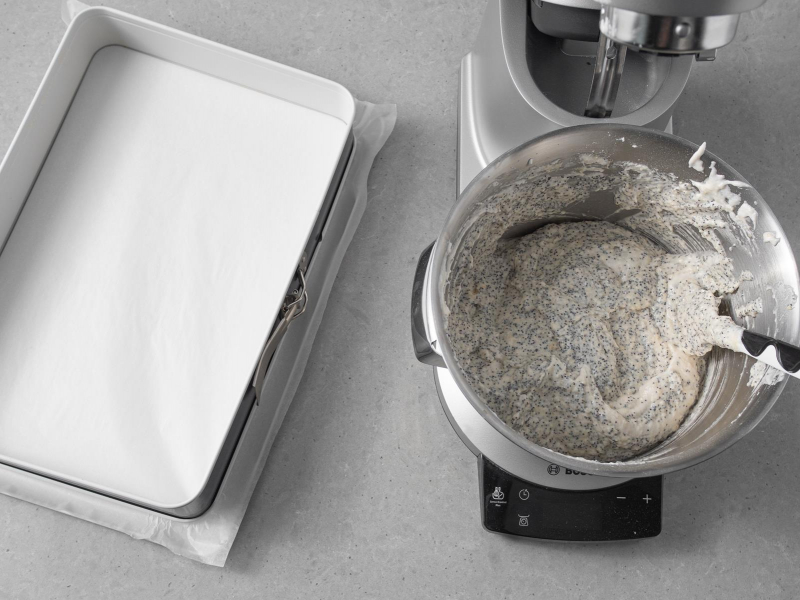 Prostokątna forma wyłożona papierem do pieczenia. Obok w misie miksera bosch masa na biszkopt makowy do makowej panienki.