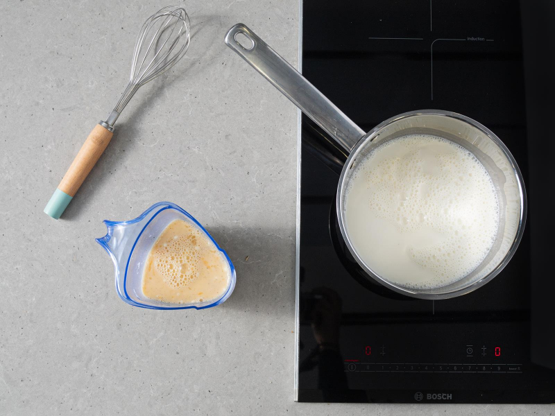 Gotujące się mleko w rondelku. Obok trzepaczka i mieszanka budyniowa w plastikowym pojemniku.