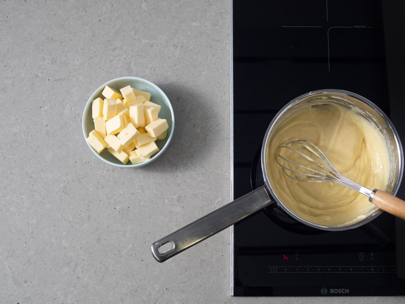 ugotowany budyń w rondelku na palniku kuchenki, obok miseczka z pokrojonym w kostkę masłem