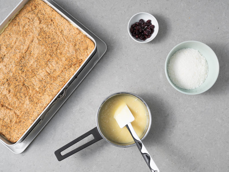 Ciasto makowa panienka w formie do pieczenia. Obok suszona żurawina w miseczce, wiórki kokosowe w drugiej miseczce oraz polewa z białej czekolady i śmietanki w rondelku..