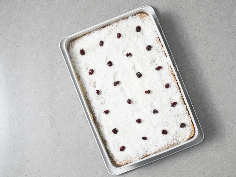 Gotowe ciasto makowa panienka polane polewą z białej czekolady, posypane wiórkami kokosowymi oraz udekorowane suszoną żurawiną namoczoną w rumie.