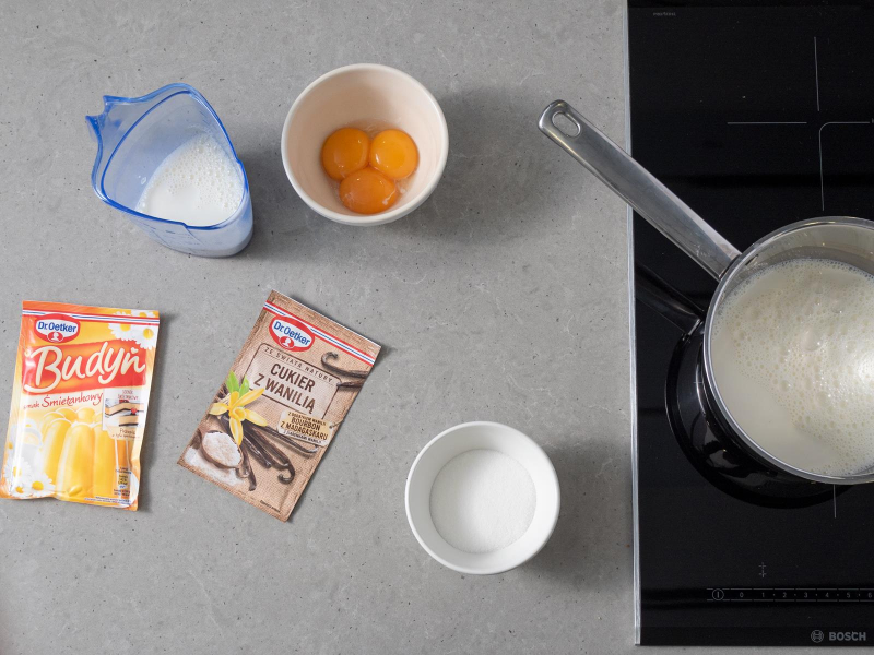 na palniku kuchenki rondelek z połową mleka, reszta mleka w pojemniku, obok miseczki z cukrem oraz żółtkami, opakowanie budyniu śmietankowego oraz opakowanie cukru z wanilią dr. oetkera