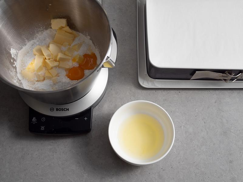 w misie robota kuchennego bosch składniki na kruche ciasto: cukier puder, mąka, mielone migdały, masło i żółtka, obok miseczka z białkami jaj oraz forma do pieczenia