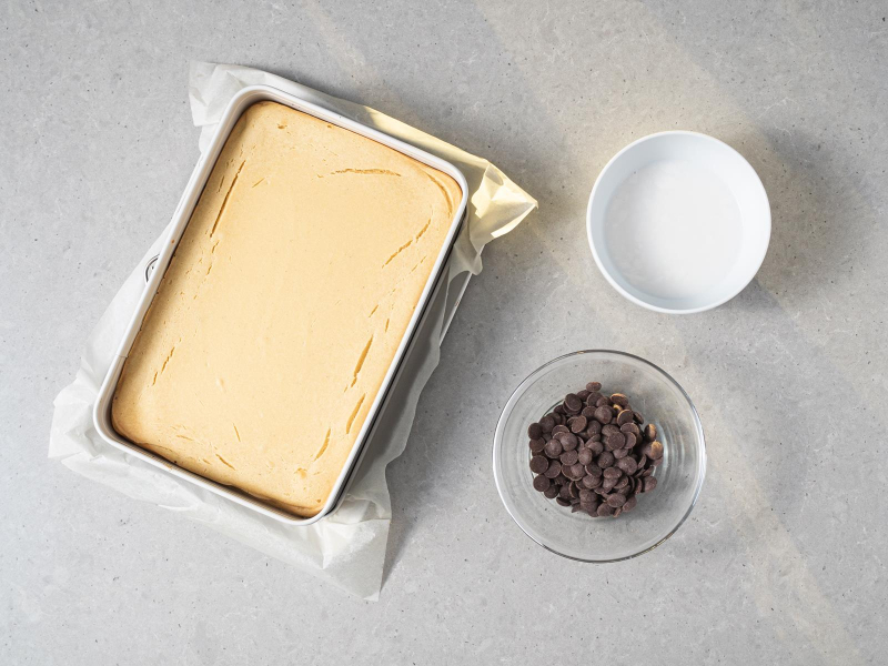 Upieczony wegański seromakowiec w formie do pieczenia. Obok czekolada gorzka w dropsach w szklanej miseczce oraz mleczko kokosowe w drugiej miseczce.