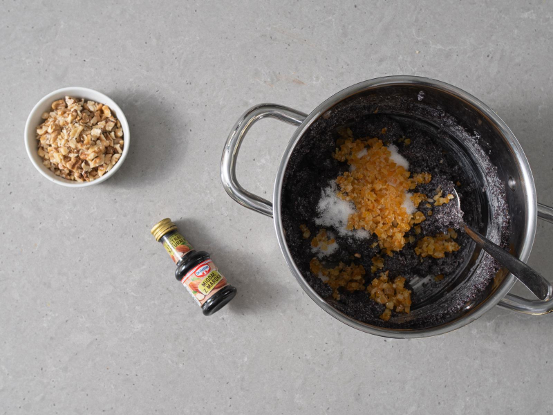 Masa makowa w garnku z dodanym cukrem i skórką pomarańczową kandyzowaną. Obok opakowanie ekstraktu migdałowego dr. oetkera oraz miseczka z posiekanymi orzechami włoskimi.