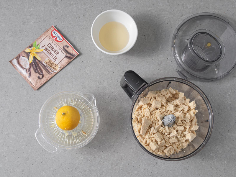 Rozdrobnione tofu w misie malaksera. Obok sok z cytryny w pojemniku do wyciskania cytryn, olej kokosowy w miseczce oraz opakowanie cukru z wanilią Dr. Oetkera.
