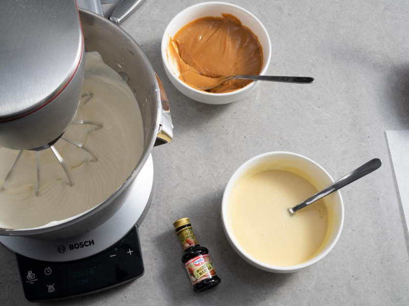 na blacie miseczka z rozpuszczoną białą czekoladą, buteleczka ekstraktu naturalnego Migdał z Marokamasą kajmakową i ubitą śmietanką