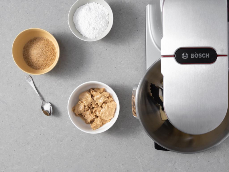 Masło orzechowe w miseczce, obok cukier puder i cukier trzcinowy.