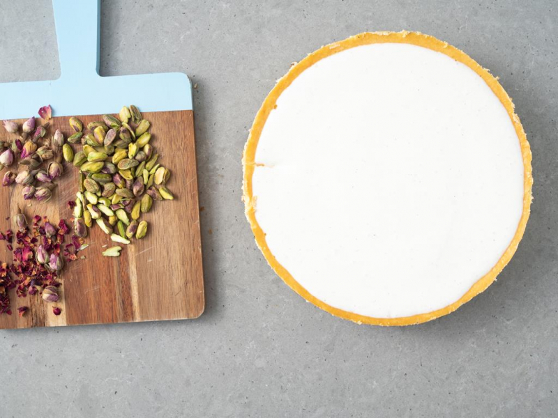 Polewa ze śmietany wylana na sernik. Obok na desce płatki suszonej róży i pistacje.