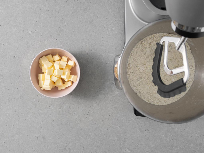 W misie robota kuchennego wymieszane suche składniki. Obok pokrojone w kostkę, zimne masło.