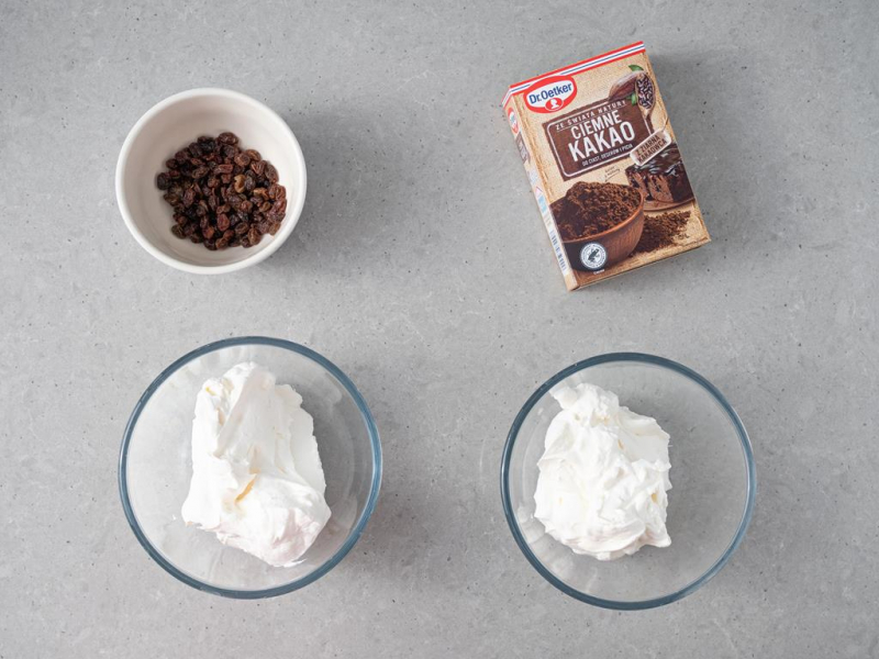 ubita śmietanka w dwóch miseczkach, obok namoczone rodzynki i kakao ciemne