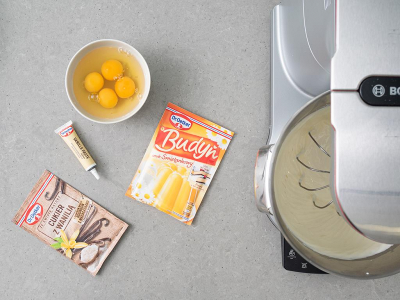 masa sernikowa w misie miksera, jajka w miseczce, opakowanie cukru z wanilią, budyń śmietankowy i pasta waniliowa