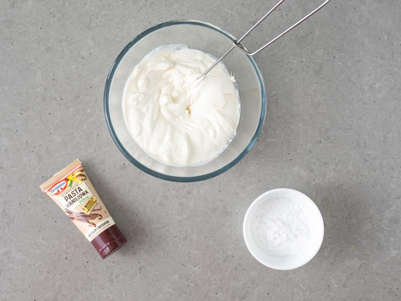 kwaśna śmietana w szklanej misce, tubka pasty waniliowe, miseczka z cukrem pudrem