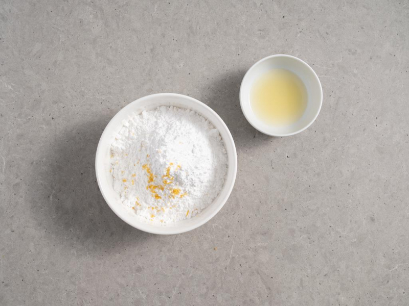 składniki na lukier cytrynowy: cukier puder i skórka z cytryny w misce, obok sok z cytryny w małej miseczce