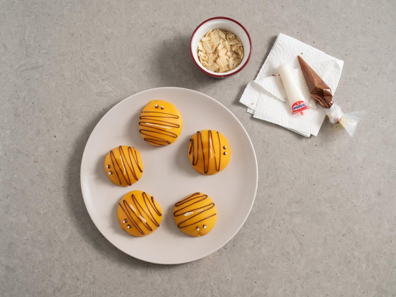 brzoskwinie udekorowane czekoladą tak, żeby przypominały pszczółki, obok rękaw cukierniczy wilton napełniony roztopioną czekoladą, biały pisak cukrowy dr. oetker oraz miseczka z płatkami migdałowymi