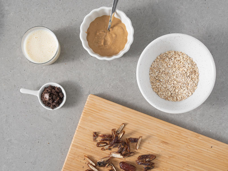 W miseczce płatki owsiane z cukrem z wanilią, proszkiem do pieczenia i solą. W miseczce masło orzechowe, napój sojowy w szklance i pokrojona gorzka czekolada. Na drewnianej desce pokrojone suszone daktyle.