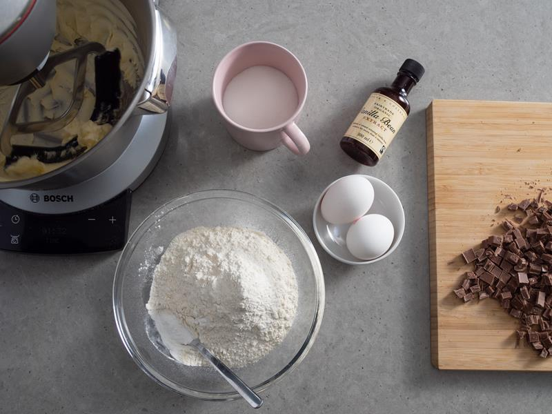 na blacie składniki do przygotowania ciasteczek piegusków: posiekana czekolada mleczna na drewnianej desce, dwa jajka w miseczce, mąka z proszkiem do pieczenia w szklanej misce, cukier w kubku, opakowanie ekstraktu waniliowego taylor and colledge oraz utarte masło w misie robota bosch