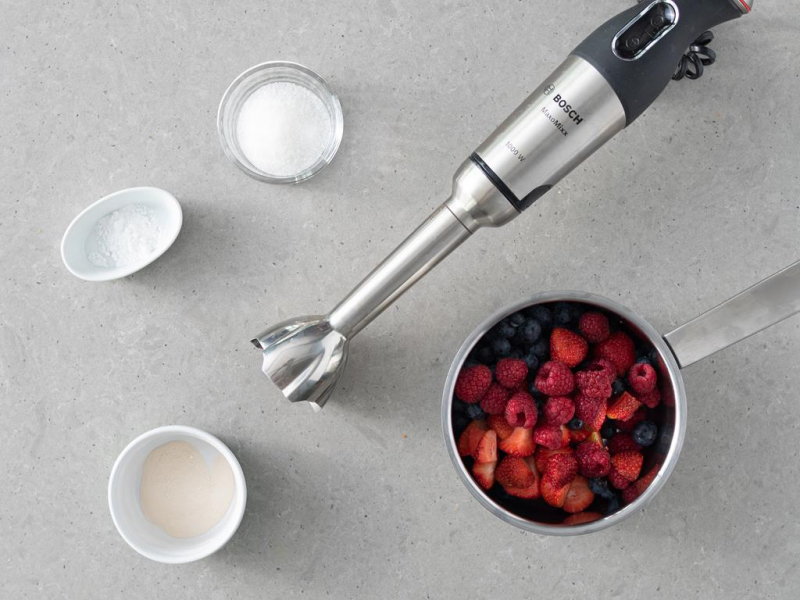 Blender ręczny firmy Bosch na blacie. Obok w garnku owoce, w miseczce agar, mąka ziemniaczana i cukier.