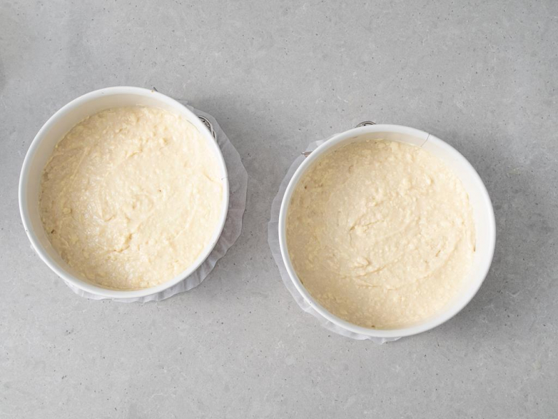 Dwie tortownice wyłożone papierem do pieczenia i wypełnione ciastem wegańskim na biszkopt.