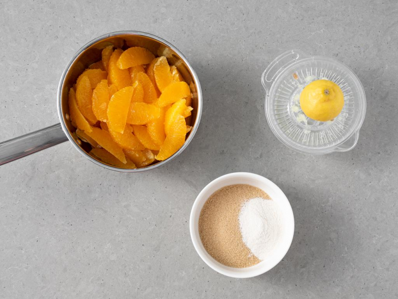 Wyfiletowane pomarańcze w garnku. Obok miseczka z cukrem trzcinowym i żelfiem oraz cytryna na wyciskarce.