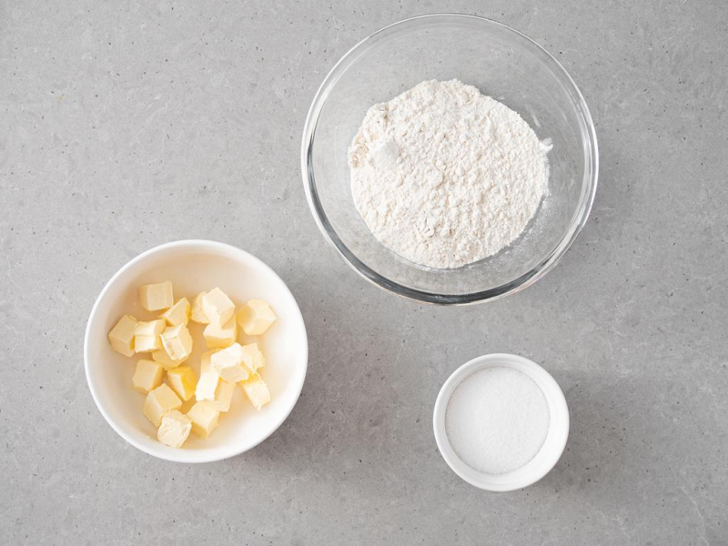 W misie mąka pszenna, oraz cukier z miseczce. Obok pokrojone w kostkę i schłodzone masło.