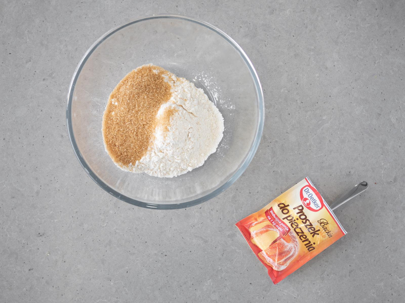 W szklanej misce mąka pszenna i brązowy cukier trzcinowy, obok opakowanie proszku do pieczenia firmy Dr. Oetker.