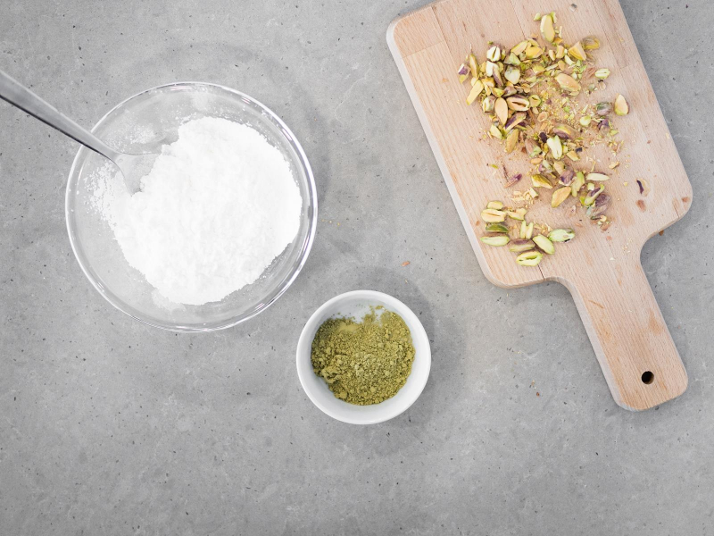 W misce cukier puder. Obok herbata matcha w małej miseczce i pokrojone pistacje na drewnianej desce.
